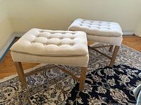 Two vintage square ottomans side by side on patterned rug, beige tufted top, wooden legs with cross braces