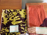Two fabric items folded on wood surface, left item is green and brown floral sheet with label, right item is reddish-orange and beige throw with label