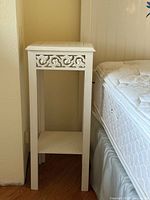 Single white vintage wooden plant stand/end table with scrollwork detail and lower shelf placed next to bed.