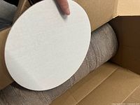 Close-up of a white, circular cardboard round being held above an open box, showing thickness and texture.