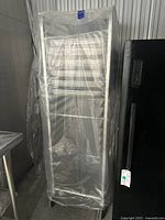 Full view of stainless steel bakery cooling rack covered in plastic wrap showing overall structure and caster wheels.