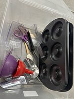 Photo showing baking items including two black donut pans, purple scissors, various scoops, funnels, and mixing bowls inside a clear plastic tote.