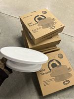 Stack of six boxed cake turntables with one taken out and shown from side view held by a gloved hand.