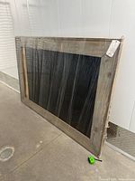 Front view of large wooden-framed chalkboard wrapped in clear protective plastic, showing grey washed finish and black chalkboard surface.