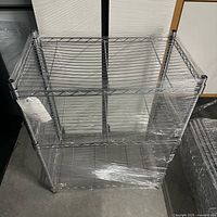 Side view of metal wire shelving unit showing three shelves and vertical posts wrapped in plastic.