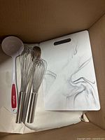 Wide view of two stainless steel handled whisks, one white ladle with red handle, and the marbled chopping board inside a cardboard box.