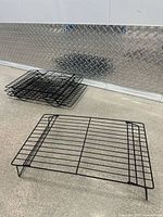 Five black wire cooling racks stacked and one set out flat showing overall shape and construction against silver diamond plate background.