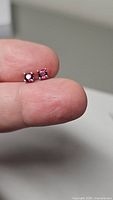 Front view of pair of blood red ruby stud earrings held between fingers showing square cut rubies with metal four-prong settings
