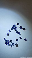Loose mixed blue sapphire gemstones with diamond cut, scattered on a white surface in circular and group formations under direct light.