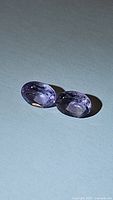 Photo showing two oval cut natural amethyst gemstones with pink-purple coloration and VS clarity, placed on a white background under direct light revealing facets and color.