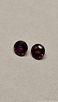 Pair of round pigeon blood red rubies on light grey background showing color and facets.