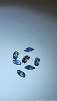 Seven marquis cut natural blue sapphires with green undertones, laid out on a neutral surface under bright light showing clarity and color.