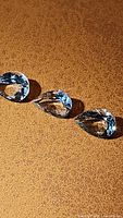 Three natural Brazilian aquamarine pear cut gemstones displayed under lighting on a textured surface, showing light blue coloration and good clarity.