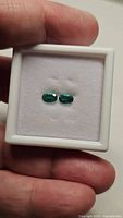 Two oval natural Columbian emeralds in white cushioned box, held by hand, showcasing intense green color and matching shape.