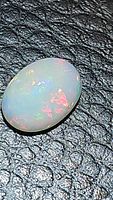 Close-up view of the oval cabochon Ethiopian opal showing the translucent white body with red and green play of color on a black textured surface.
