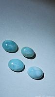 Four oval natural Larimar cabochon gemstones on a grey background, showcasing their glossy finish and blue-white marbling.
