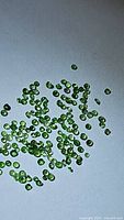 Close-up photo showing numerous very small bright green tsavorite garnet stones scattered on a white surface, highlighting their size and diamond cut faceting.
