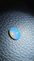 Close-up photo of a natural oval opal cabochon displaying blue, yellow, and green flashes on black textured background.