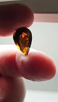 Photo shows the pear cut cognac orange tourmaline gemstone held between thumb and forefinger, backlit to show color and facets.