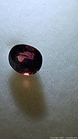 Close-up photo of the untreated Winza ruby on a flat surface under natural light, showing the oval cut and deep dark red color with highlights.