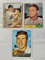 Three vintage baseball cards are shown together on a flat surface. Front images of players Stan Musial, Brooks Robinson and Billy Martin are clearly visible. The cards exhibit wear such as rounded corners and creases.
