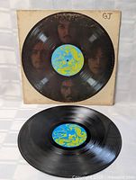 Black vinyl LP record with blue and yellow label on a photo background of band members' faces on the sleeve, the sleeve shows typical wear and some discoloration.