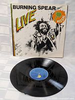Front view of Burning Spear LIVE reggae vinyl vinyl record with cover showing colorful title, artist and reggae sticker illustration, alongside the black vinyl disc with blue label.