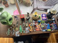 Overhead view of all alien-themed items including loose figurines, packaged figures, art prints, alien head container, UFO playset, and space lamp.