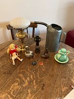 Photo showing entire collection of antique collectibles on wooden table including lamp, doll, oil can, candle holder, small green floral china set, mini pitcher, wooden nickel, wooden duck figurine, belt buckle, and coin.