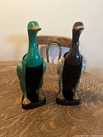 Front view of two duck-shaped brushes on a wooden table, showing green and black painted ducks with gold accents and bristle brush bodies.