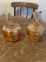 Two amber glass containers on a wooden table, each with an animal figurine lid - dog and deer.