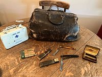 Overall view showing the leather doctor's bag, white metal first aid kit, three straight razors with cases, and packaging on a wooden table.