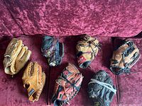 Seven leather baseball gloves with various colors and brands on a red velvet couch
