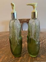 Front view of two glass corn-shaped dispensers with white pumps on a wooden table.