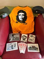 Shows a red armchair with an orange t-shirt featuring Che Guevara image, two revolutionary style caps on each armrest and seven assorted black and white and color postcards and photos on the seat.