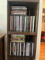 Two wooden shelves filled with stacks of CDs. Titles and artists appear diverse, in jewel cases with minor wear. CDs are organized in rows within shelves.