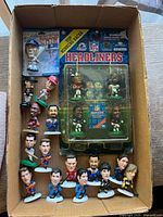 Photo showing all loose and packaged sports mini figurines in an open box, including football, basketball, baseball, hockey figures, and sealed NFL Headliners running backs set.