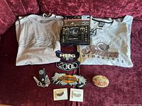 Two Indian Motorcycle t-shirts laid out on fabric surface with patches, pins, wallet chain, small tin, and bike bag.