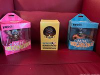 Three Muchacho bobblehead figures in their original packaging sitting side by side on a red background: one in a pink box, one in a yellow box, one in a blue box.