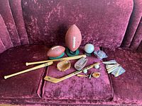 Various small sports-themed novelty items arranged on couch, showing two football snack bowls, miniature bats, hockey stick, baseball gloves, and a ceramic baseball piggy bank.