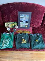 Overview of entire Oakland Athletics memorabilia set including shirts, patch, license plate, framed photo, money clip, and baseball laid out on a red chair.