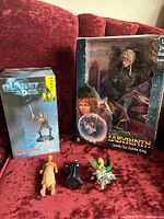 Front view of all items on red fabric couch: boxed Jareth The Goblin King figure, boxed Planet of the Apes Thade figure, and three small Disney mini figurines unboxed.