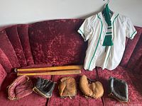 Photo showing two vintage baseball jerseys, two wooden bats, five vintage gloves laid out on red couch