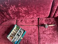 Full view of fishing rod alongside an open tackle box showing assorted fishing lures and tools inside compartments on a red fabric couch.