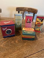 Five vintage Fossil branded tins displayed on table, showing wide variety of designs and colors including one red tin with built-in clock face.