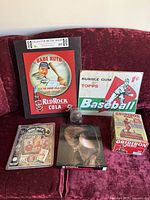 Collection of vintage sports memorabilia on red couch including Babe Ruth metal sign, Topps baseball gum sign, Hockey Immortals card set and book, leather baseball, and two sports books.