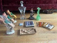Photo showing lady figurines, glass male torso decanter, green dress figurine, black woman figurine, ashtray with reclining lady, and illustrated trading cards laid out on table.