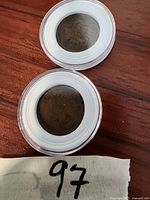 Two Indian Head pennies dated 1889 and 1908 in clear protective capsules on wooden surface with handwritten paper showing number 97.