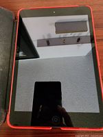 Front view of Apple iPad Mini tablet in red protective case on a wooden surface showing touchscreen and home button.