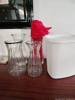 Four ribbed glass vases of various sizes on wooden surface, red cloth inside one vase, white plastic trash can next to vases